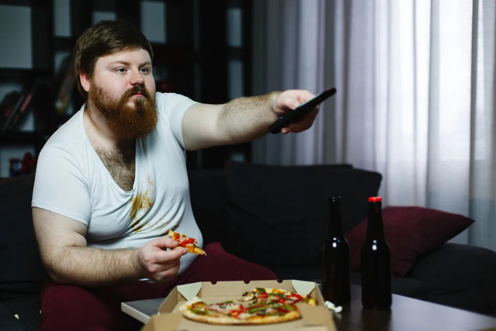 Overweight man eating pizza and drinking beer, illustrating lifestyle factors related to Obesity in Men.