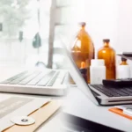 Two healthcare professionals review medical documents at a desk with a laptop, clipboard, and medicinal bottles nearby.