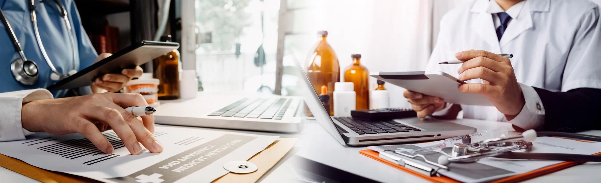Two healthcare professionals review medical documents at a desk with a laptop, clipboard, and medicinal bottles nearby.